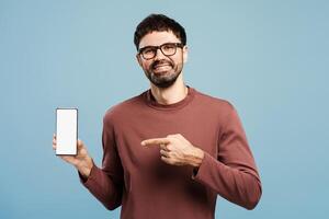 Smiling Hispanic man holding mobile phone with empty screen, pointing finger on mockup, copy space photo