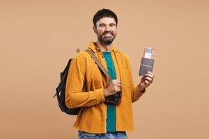 Smiling, attractive bearded man, traveler holding backpack and passport with ticket, looking away photo