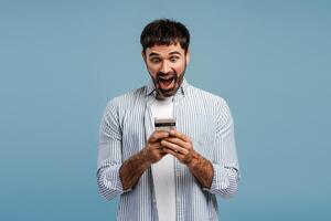 Excited, bearded man holding mobile phone, using mobile win money, isolated on blue background photo