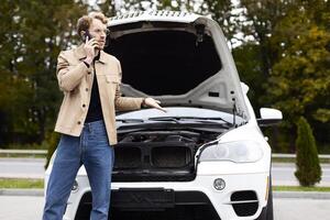 Confused driver calling mechanic on mobile phone near broken car with open hood photo
