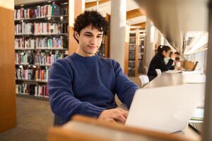 Young man working on laptop sitting at desk in busy public library photo