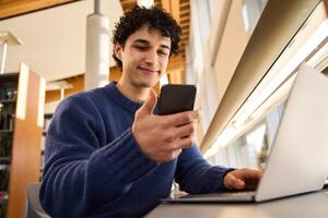 Young man using smartphone while working on laptop in library photo