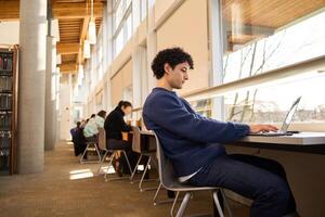 Focused college student working on laptop in busy university library photo