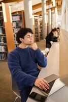 Thoughtful student working on laptop in busy college library photo