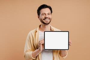 Smiling, bearded man showing digital tablet with empty screen on beige background looking at camera photo