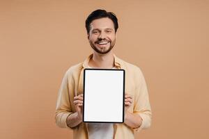 Happy man showing digital tablet with empty screen standing over beige background looking at camera photo