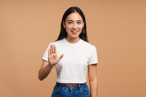Young, beautiful Asian woman smiling and showing stop gesture with hand on beige background photo