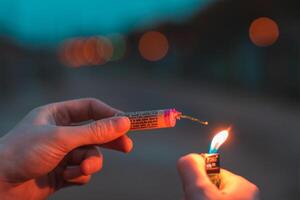 Young Man Lighting Up Firecracker in his Hand Outdoors in Evening. Guy Getting Ready for New Year Fun with Fireworks or Pyrotechnic Products - CloseUp Shot, Rear View photo