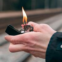 Setting Fire to the Firecracker. Man in Black Clothes Lighting Up the Petard. Firing Up the Pyrotechnics with a Black Gasoline Lighter Outdoors photo