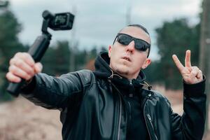 Blogger in Sunglasses Making Selfie or Streaming at the Pine Forest Using Action Camera with Gimbal Camera Stabilizer. Handsome Man in Black Clothes Making Photo