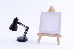 Wooden Easel Miniature with Blank White Square Canvas for Artists and Painters - Mockup. Mini Wooden Stand with Clean Artboard and Small Black Table Lamp on White Background, Copy Space photo