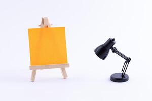 Wooden Easel Miniature with Blank Colored Square Canvas or Memo Paper - Mockup. Mini Wooden Stand with Clean Artboard and Small Black Table Lamp on White Background, Copy Space photo
