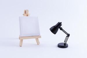 Wooden Easel Miniature with Blank White Square Canvas for Artists and Painters - Mockup. Mini Wooden Stand with Clean Artboard and Small Black Table Lamp on White Background, Copy Space photo