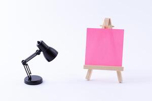 Wooden Easel Miniature with Blank Colored Square Canvas or Memo Paper - Mockup. Mini Wooden Stand with Clean Artboard and Small Black Table Lamp on White Background, Copy Space photo