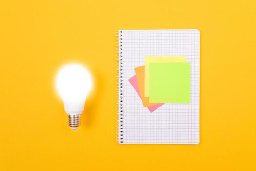 An Idea Concept - Glowing Power Saving Lamp and A Notepad with Sticky Notes Lying on Yellow Table. Top View, Flat Lay, Blank Template photo