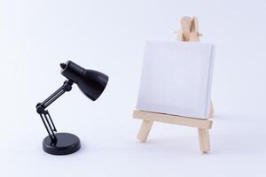 Wooden Easel Miniature with Blank White Square Canvas for Artists and Painters - Mockup. Mini Wooden Stand with Clean Artboard and Small Black Table Lamp on White Background, Copy Space photo