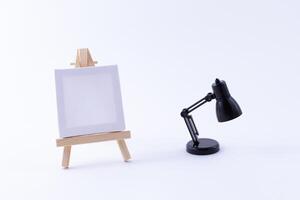 Wooden Easel Miniature with Blank White Square Canvas for Artists and Painters - Mockup. Mini Wooden Stand with Clean Artboard and Small Black Table Lamp on White Background, Copy Space photo