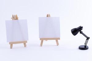 Two Wooden Easels Miniature with Blank White Square Canvas for Artists and Painters - Mockup. Mini Wooden Stands with Clean Artboard and Small Black Table Lamp on White Background, Copy Space photo