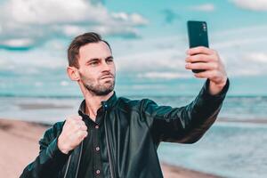 Young Blogger Making Selfie or Streaming at the Beach Using Black Smartphone. Handsome Man in Black Clothes Making Photo Against the Sea at Cloudy Day