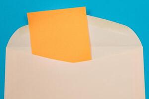 Beige Envelope with Blank Orange Sheet of Paper Inside, Lying on Blue Background - Mock Up with Copy Space. Receiving Mail or Notice, Sending Postcard - Top View, Flat Lay photo