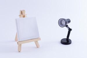 Wooden Easel Miniature with Blank White Square Canvas for Artists and Painters - Mockup. Mini Wooden Stand with Clean Artboard and Small Black Table Lamp on White Background, Copy Space photo