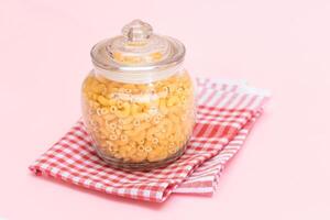 Uncooked Chifferi Rigati Pasta in Glass Jar on Checkered Red Towel on Pink Background. Fat and Unhealthy Food. Scattered Classic Dry Macaroni. Italian Culture and Cuisine. Raw Pasta photo