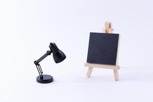 Wooden Easel Miniature with Blank Black Square Canvas for Artists and Painters - Mockup. Mini Wooden Stand with Clean Artboard and Small Black Table Lamp on White Background, Copy Space photo