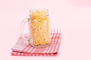 Uncooked Chifferi Rigati Pasta in Glass Jar on Checkered Red Towel on Pink Background. Fat and Unhealthy Food. Scattered Classic Dry Macaroni. Italian Culture and Cuisine. Raw Pasta photo