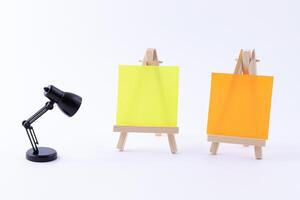 Two Wooden Easels Miniature with Blank Colored Square Canvas or Memo Paper - Mockup. Mini Wooden Stand with Clean Artboard and Small Black Table Lamp on White Background, Copy Space photo