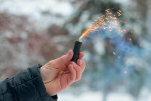 Burning Firecracker in a Hand. Guy Holding a Petard Outdoors in Winter at Daytime. Loud and Dangerous New Year's Entertainment. Hooliganism with Pyrotechnics. Noise of Firecrackers in Public Places photo