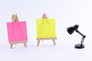 Two Wooden Easels Miniature with Blank Colored Square Canvas or Memo Paper - Mockup. Mini Wooden Stand with Clean Artboard and Small Black Table Lamp on White Background, Copy Space photo