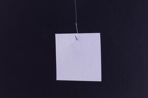 Mockup of a Blank White Memo Paper with Copy Space Hanging on a Fishing Hook Against the Black Background. Reminder or To Do List. Sticky Note Template photo