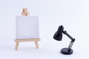 Wooden Easel Miniature with Blank White Square Canvas for Artists and Painters - Mockup. Mini Wooden Stand with Clean Artboard and Small Black Table Lamp on White Background, Copy Space photo