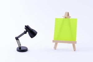 Wooden Easel Miniature with Blank Colored Square Canvas or Memo Paper - Mockup. Mini Wooden Stand with Clean Artboard and Small Black Table Lamp on White Background, Copy Space photo