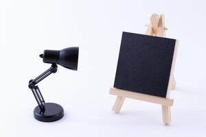 Wooden Easel Miniature with Blank Black Square Canvas for Artists and Painters - Mockup. Mini Wooden Stand with Clean Artboard and Small Black Table Lamp on White Background, Copy Space photo
