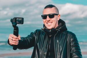 Smiling Young Male Blogger in Sunglasses Making Selfie or Streaming at the Beach Using Action Camera with Gimbal Camera Stabilizer. Hipster in Black Clothes Making Photo Against the Sea