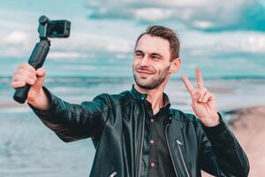 Smiling Young Blogger Making Selfie or Streaming at the Beach Using Action Camera with Gimbal Camera Stabilizer. Man in Black Clothes Making Photo Showing Peace