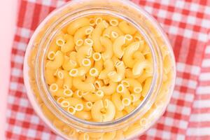 Uncooked Chifferi Rigati Pasta in Glass Jar on Checkered Red Towel on Pink Background. Fat and Unhealthy Food. Scattered Classic Dry Macaroni. Italian Culture and Cuisine. Raw Pasta photo