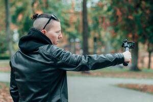 Blogger in Black Clothes and Sunglasses Making Using Action Camera with Gimbal Camera Stabilizer in Autumn Park photo