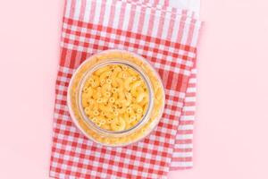 Uncooked Chifferi Rigati Pasta in Glass Jar on Checkered Red Towel on Pink Background. Fat and Unhealthy Food. Scattered Classic Dry Macaroni. Italian Culture and Cuisine. Raw Pasta photo