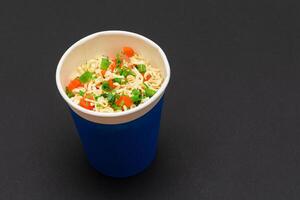 Prepared Instant Noodles with Carrot and Greens in Blue Cup on Black Background. Quick Lunch or Unhealthy Fast Food photo