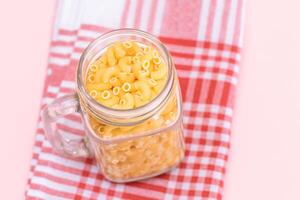 Uncooked Chifferi Rigati Pasta in Glass Jar on Checkered Red Towel on Pink Background. Fat and Unhealthy Food. Scattered Classic Dry Macaroni. Italian Culture and Cuisine. Raw Pasta photo