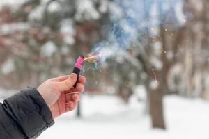 Burning Firecracker in a Hand. Guy Holding a Petard Outdoors in Winter at Daytime. Loud and Dangerous New Year's Entertainment. Hooliganism with Pyrotechnics. Noise of Firecrackers in Public Places photo