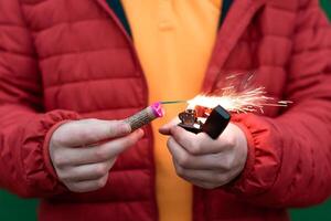 Man in Red Jacked Lighting Up Firecracker in his Hand Using Gasoline Lighter. Guy Getting Ready for New Year Fun with Fireworks or Pyrotechnic Products photo