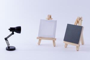 Two Wooden Easels Miniature with Blank Black and White Square Canvas for Artists and Painters - Mockup. Mini Wooden Stand with Clean Artboard and Small Black Table Lamp on White Background, Copy Space photo