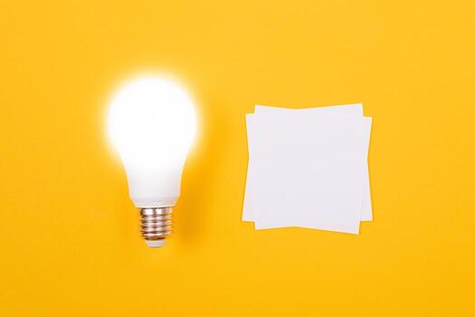 Glowing Power Saving Lamp with White Note Paper Lying on Yellow Table, Blank Template with Copy Space - Top View, Flat Lay photo