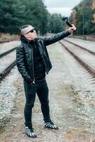 Blogger in Sunglasses Making Selfie or Streaming at the Pine Forest Using Action Camera with Gimbal Camera Stabilizer. Handsome Man in Black Clothes Making Photo