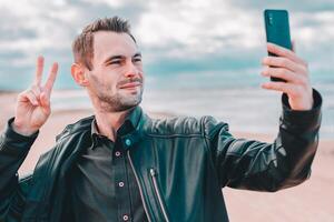Young Blogger Making Selfie or Streaming at the Beach Using Black Smartphone. Handsome Man in Black Clothes Making Photo Against the Sea at Cloudy Day
