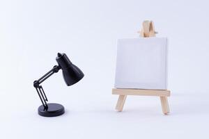 Wooden Easel Miniature with Blank White Square Canvas for Artists and Painters - Mockup. Mini Wooden Stand with Clean Artboard and Small Black Table Lamp on White Background, Copy Space photo