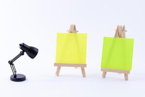 Two Wooden Easels Miniature with Blank Colored Square Canvas or Memo Paper - Mockup. Mini Wooden Stand with Clean Artboard and Small Black Table Lamp on White Background, Copy Space photo